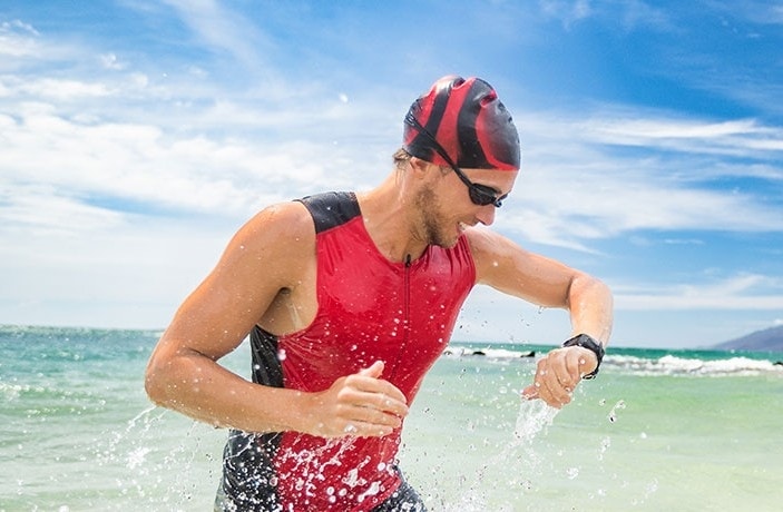 一位身穿红色铁人三项运动服、佩戴运动手表的运动员，正从浪花飞溅的海水中跑向岸边。