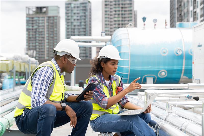 在工厂环境中，两名头戴红色安全帽、身穿黄色安全背心的工人正在操作和检查一台大型工业机器人。