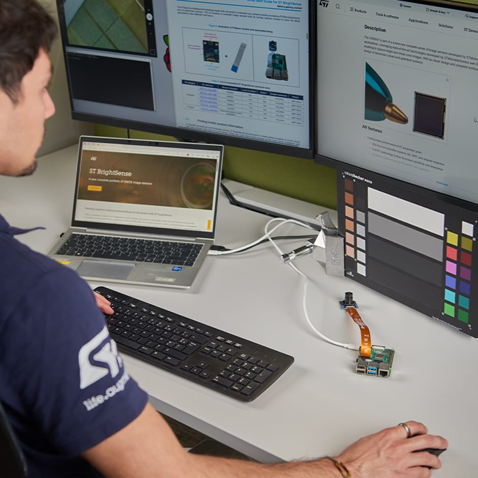 一名身着意法半导体马球衫的男子正在办公桌前工作，桌上有一台笔记本电脑、三台显示技术内容的显示器、一个键盘以及一块电子电路板。