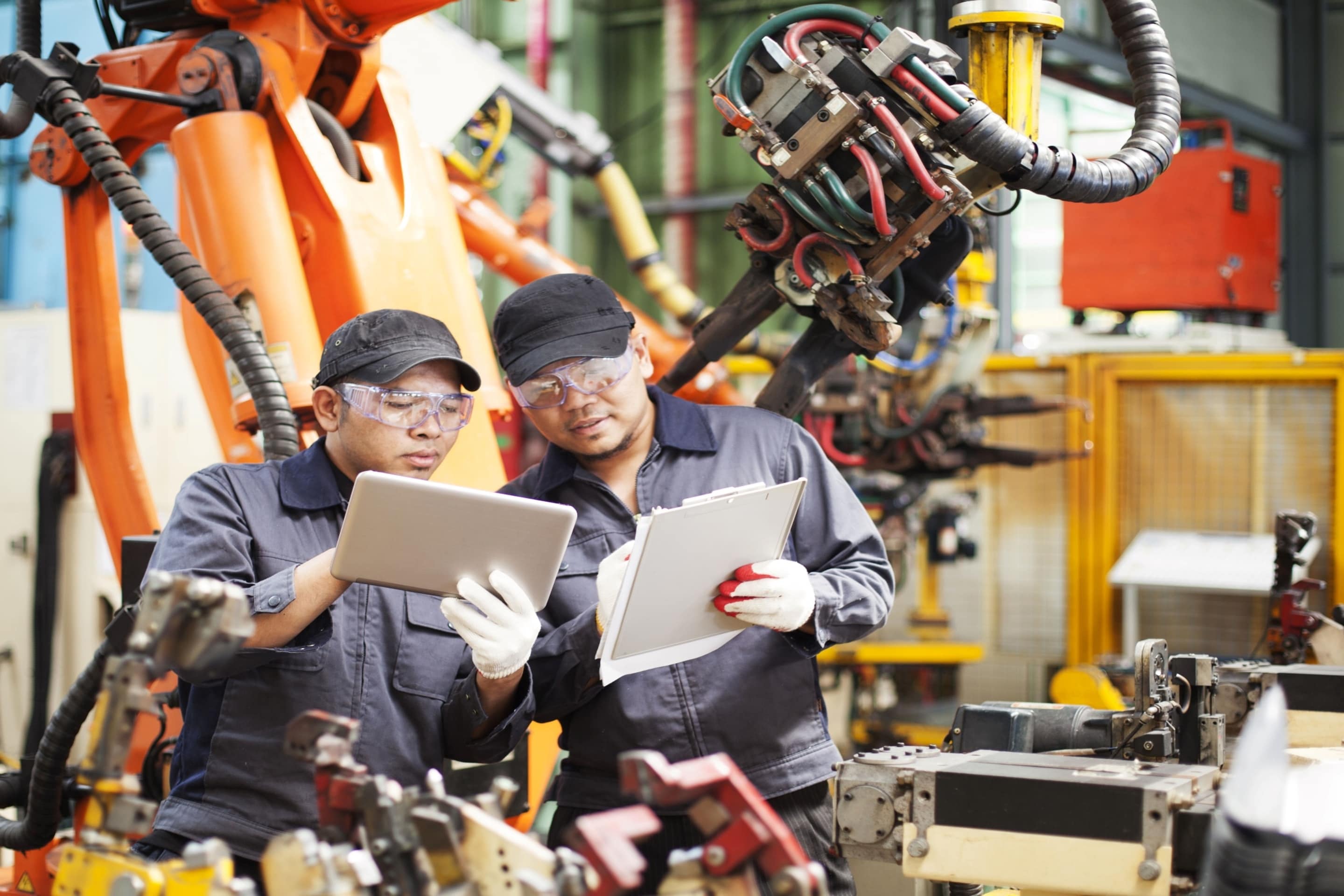 Two workers in a factory setting reviewing information on a tablet and clipboard, surrounded by industrial robotic arms and machinery.