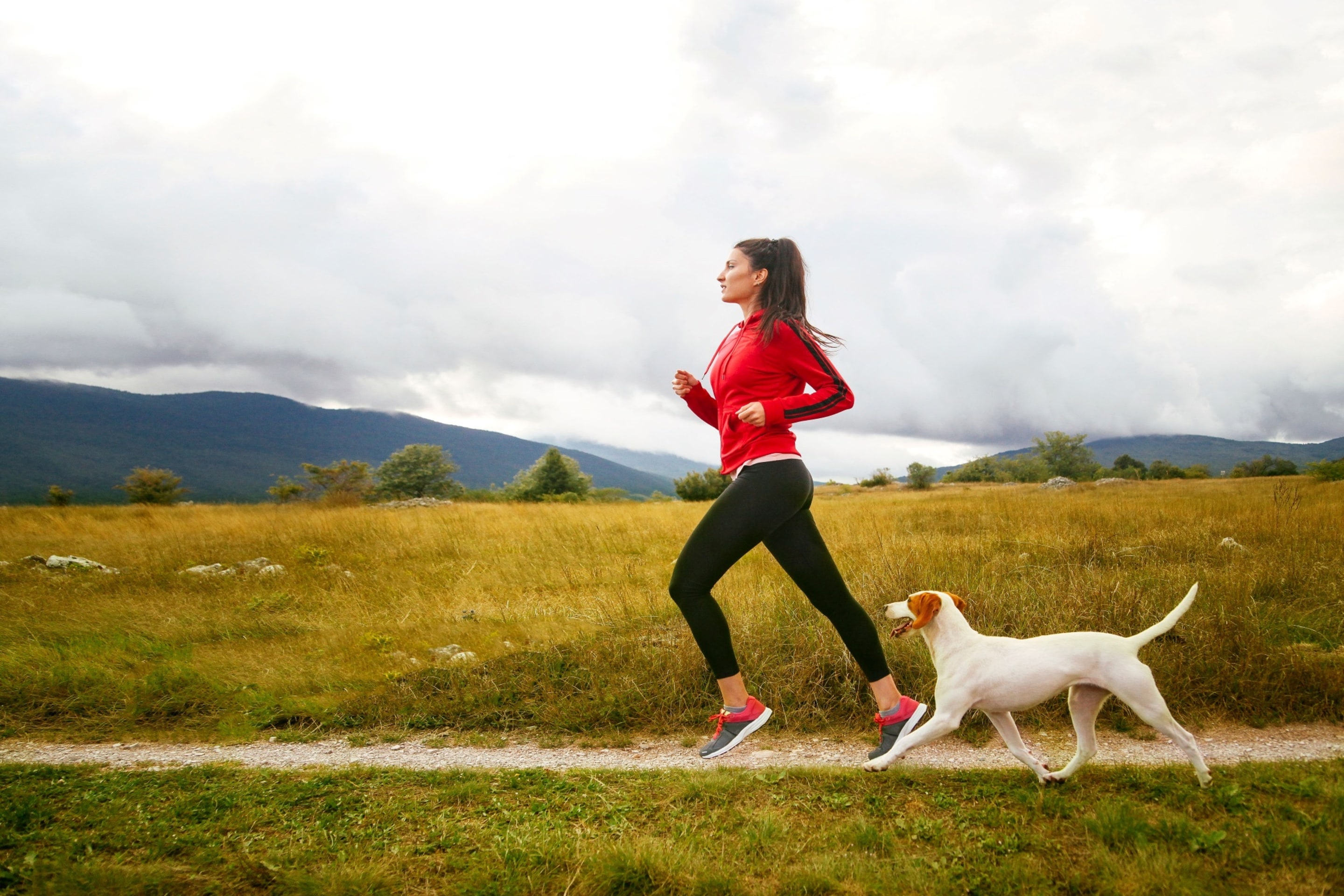 Woman jogging on a grassy trail in a natural landscape with mountains and cloudy sky, accompanied by a white dog.