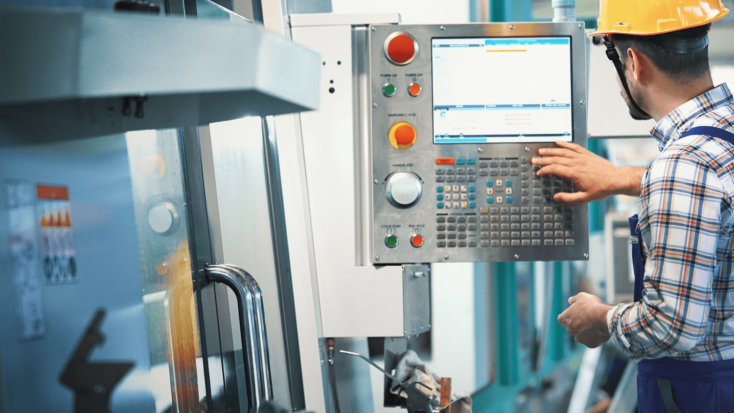 Man wearing a yellow safety helmet operating a CNC machine control panel in a factory.